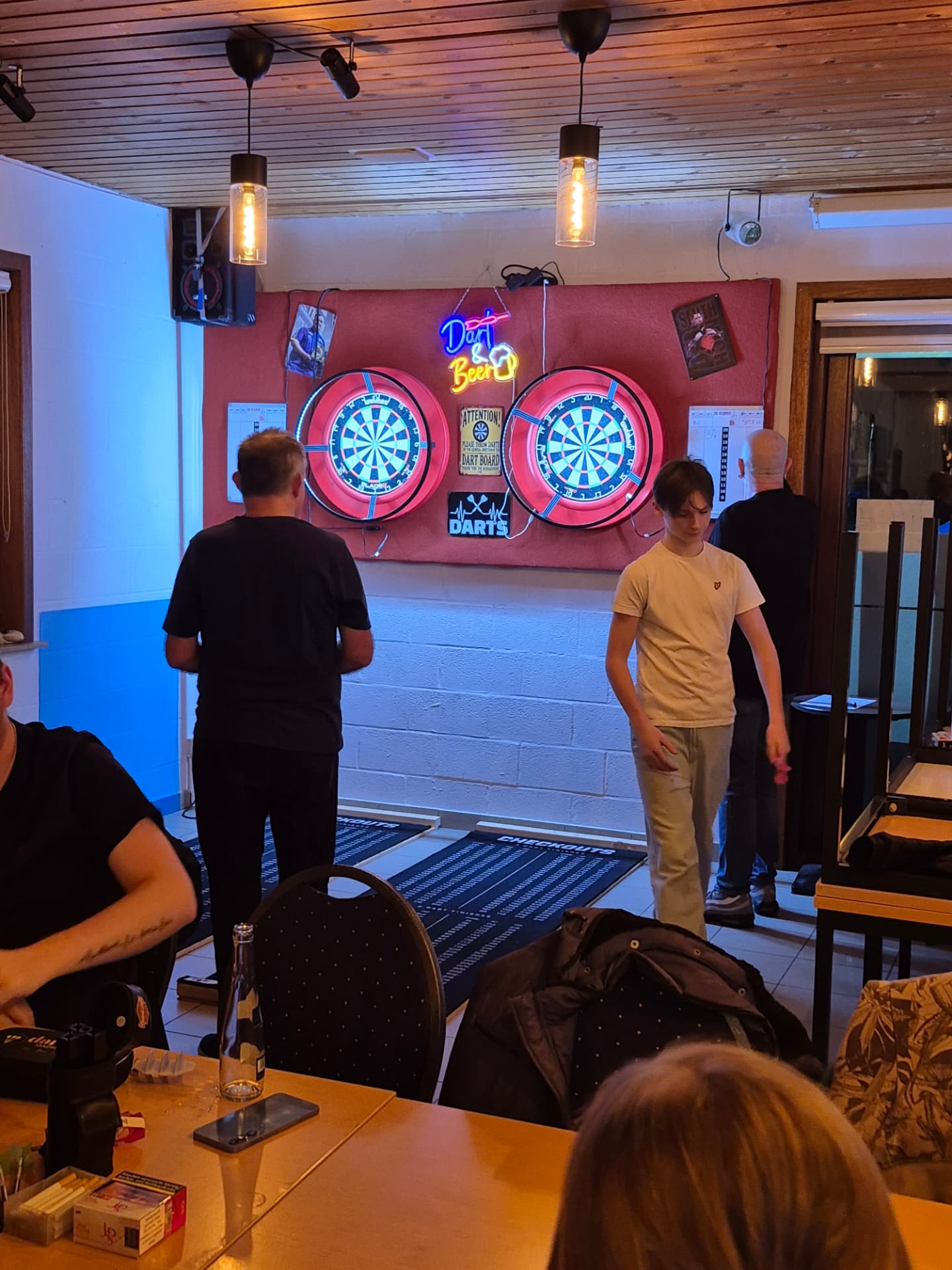 Spelers aan de dartborden met de neon 'Dart & Beer' tijdens de clubavond